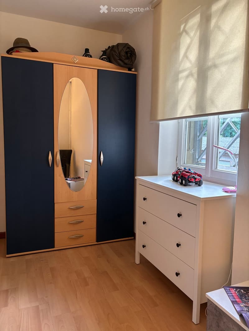 Blue wardrobe with mirror, white chest of drawers, toy car, hat, and lamp on window sill