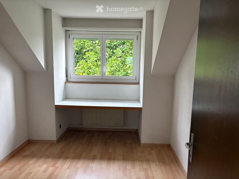 Empty room with wooden flooring, white walls, a window with a view of greenery, and a built-in bench beneath the window.