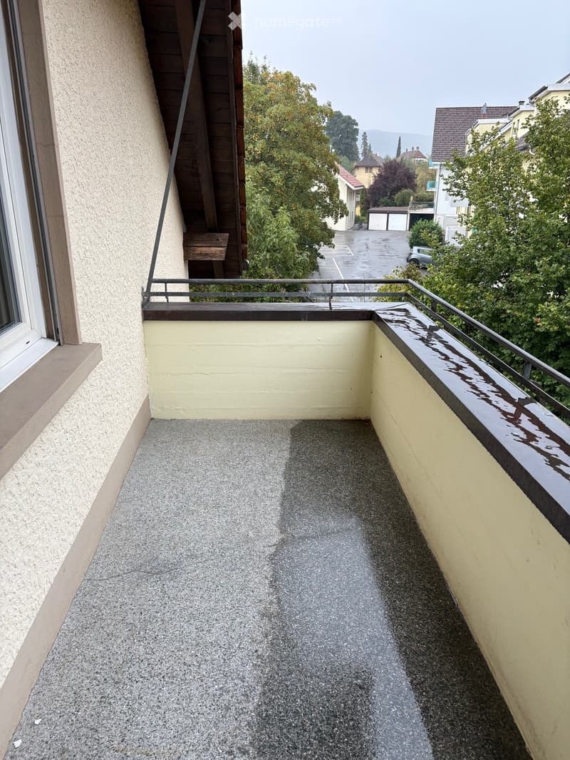 Balcony with grey concrete floor, beige walls, metal railing, and a view of the surrounding neighborhood with trees and buildings.