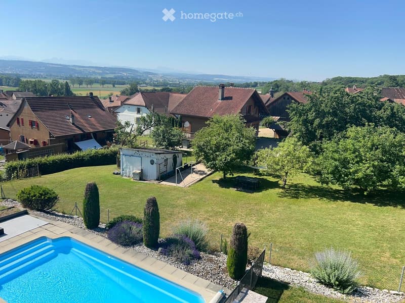 Aerial view of a residential area with multiple houses, a swimming pool, a lawn, trees, and a scenic countryside backdrop.