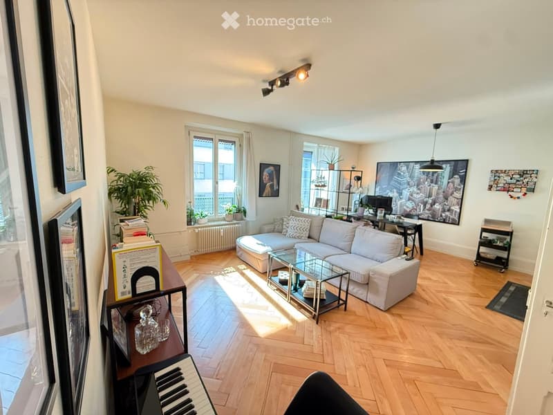 Bright living room with wooden flooring, large windows, a white sectional sofa, a glass coffee table, a piano, and decorative artwork.