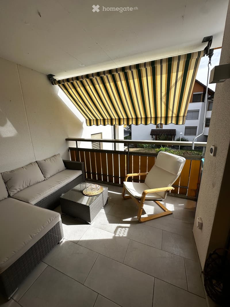 Balcony with a striped awning, a wicker sofa, a wooden chair, and a small table with a cushion.