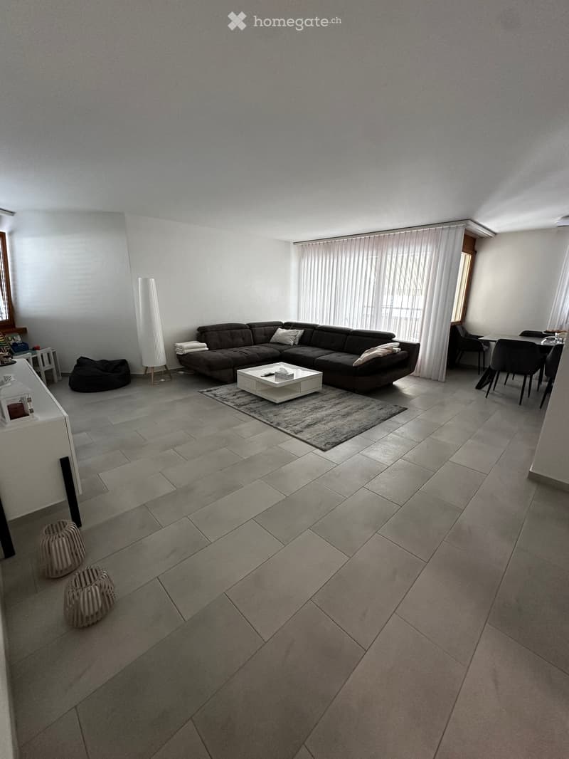 Modern living room with grey sofa, white coffee table, dining area with chairs and table, and decorative elements including a floor lamp and woven stools.