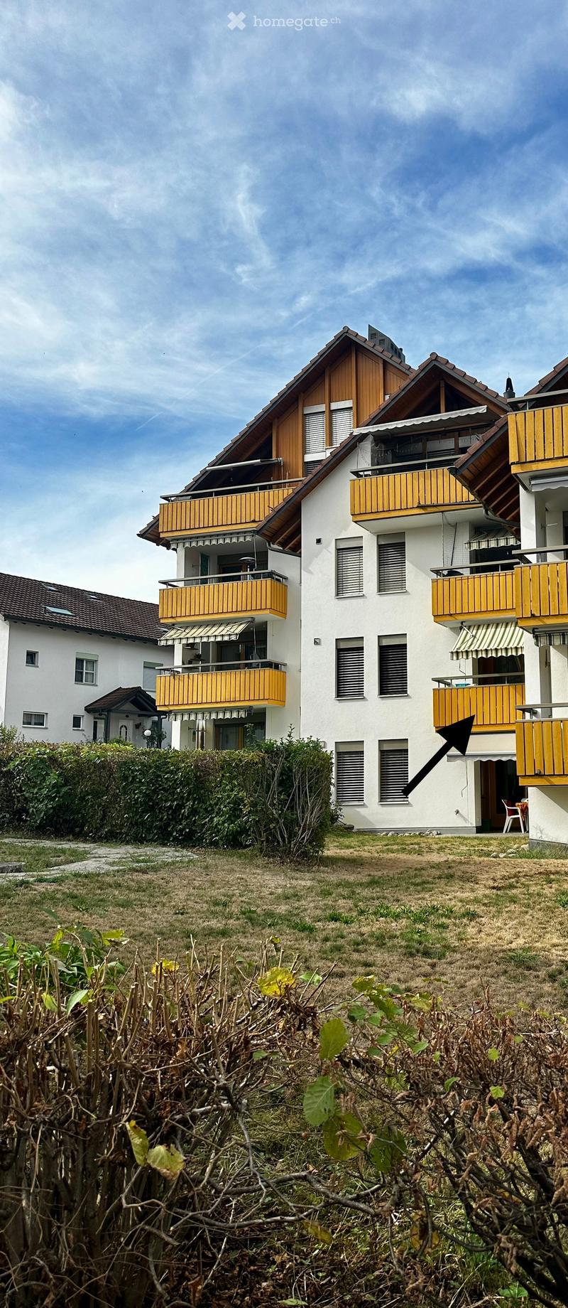 A multi-story residential building with white walls, wooden balconies, and a sloped roof. The building has multiple windows and a small garden area in front.