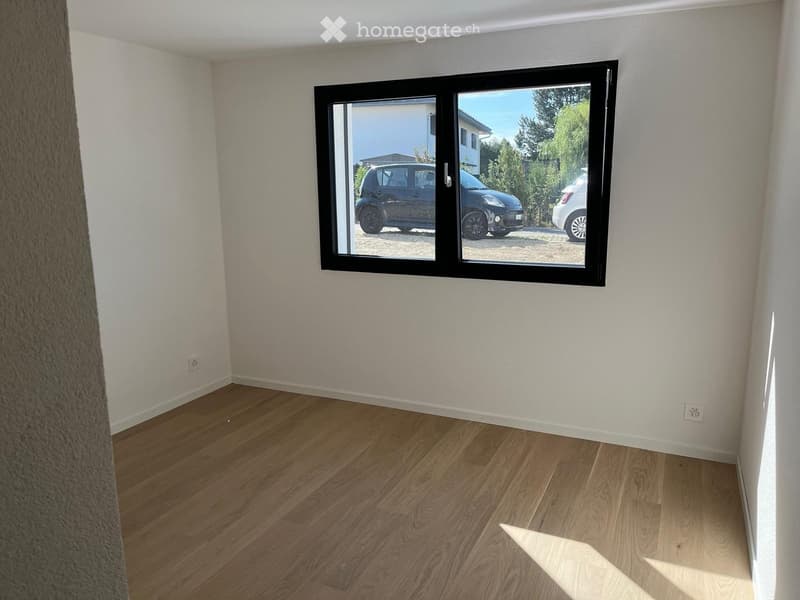 Empty room with wooden flooring and white walls, featuring a large window with a view of parked cars and greenery outside.