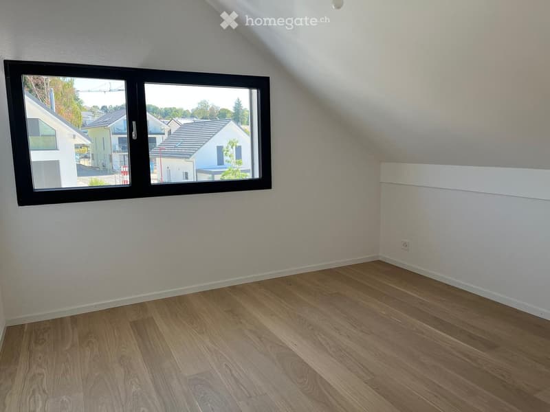 Empty attic room with wooden flooring, white walls, and a large window offering a view of the neighborhood.