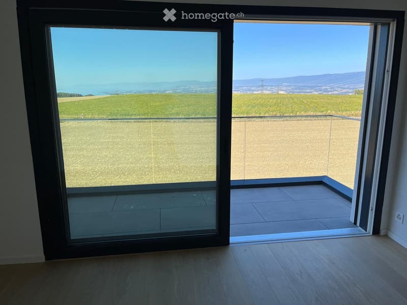 Open sliding glass doors lead to a tiled balcony with a panoramic view of fields and distant hills.