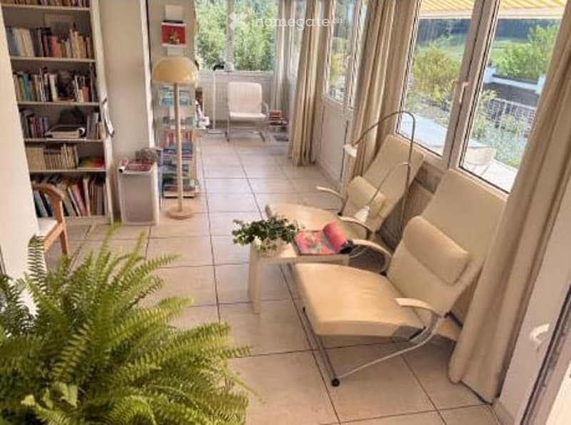 Bright living room with large windows and a view, featuring a sofa, chairs, a small table, bookshelf, and a floor lamp.