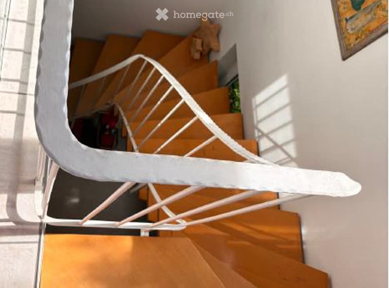 Spiral staircase with wooden steps and white handrail, leading to an upper level. The walls are white, and there is a small plant visible on the landing.