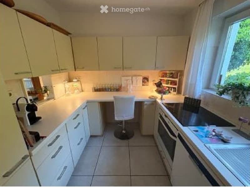 Bright kitchen with white cabinets, tiled floor, and a large window. Includes a coffee maker, fridge, sink, and a small table with chairs.