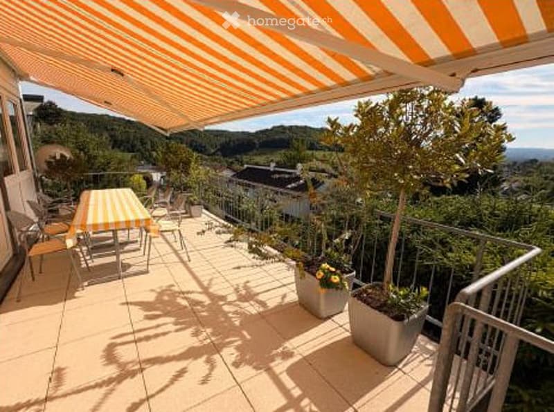 Balcony with striped awning, outdoor table and chairs, potted plants, scenic hillside view.