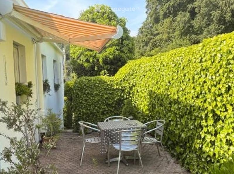 A small patio area with a table and chairs, surrounded by a neatly trimmed hedge and a house with a striped awning.