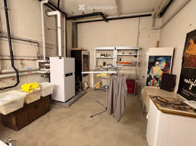 Empty utility room with exposed pipes, a water heater, shelving units, and a small table with a poster on it.