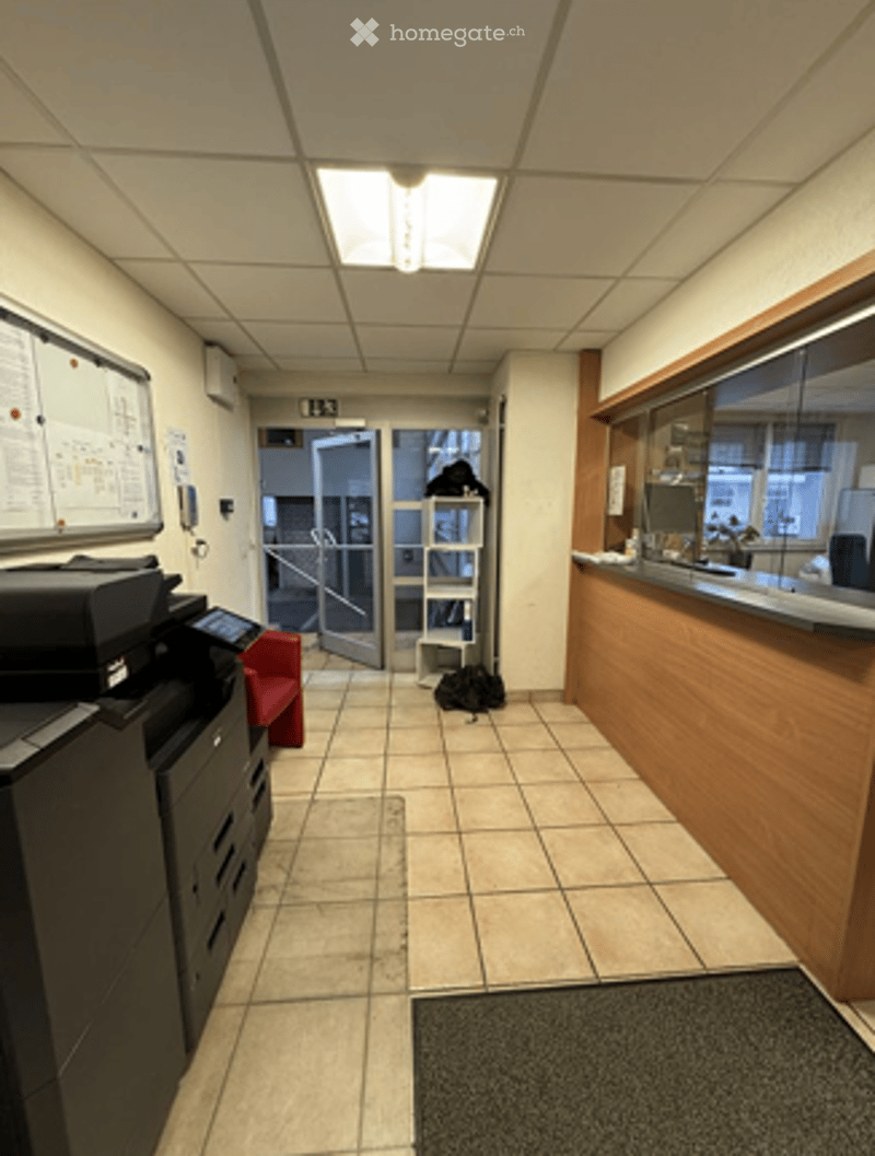 Empty hallway with tiled floor, white walls, and a reception area visible at the end. There is a printer, a chair, and a small table with items on it.