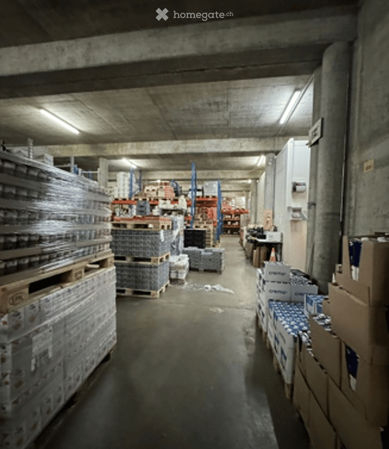 Warehouse space with concrete floors and ceilings, filled with pallets of boxes and shelves stocked with various items. Worktable and storage units visible along the sides.