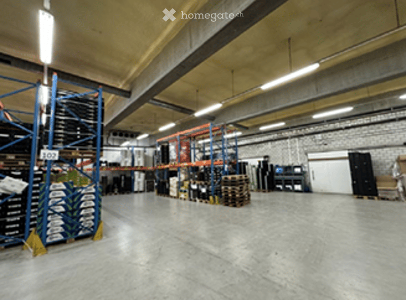 Large industrial-style warehouse with high ceilings, exposed beams, and bright lighting. Shelves, pallets, and storage units are visible, along with a computer setup in the background.