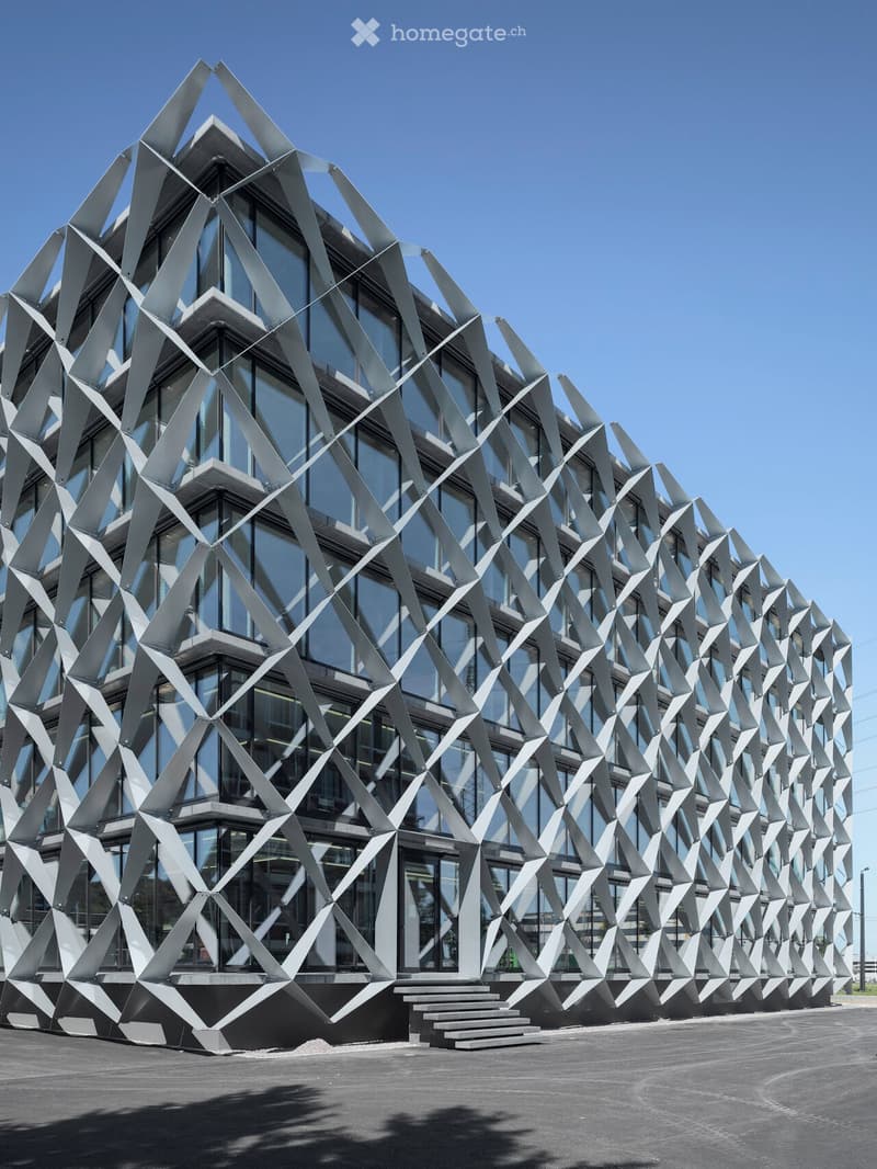 Modern building with a geometric, angular metal facade and large glass windows, featuring a raised ground floor and steps leading to the entrance.
