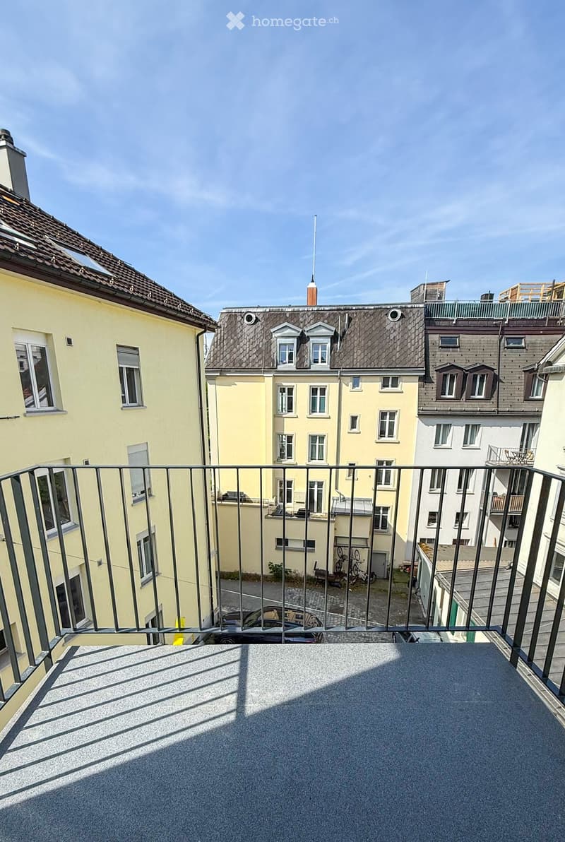 Balcony overlooking a cityscape with multiple residential buildings, metal railing, and parked cars below.