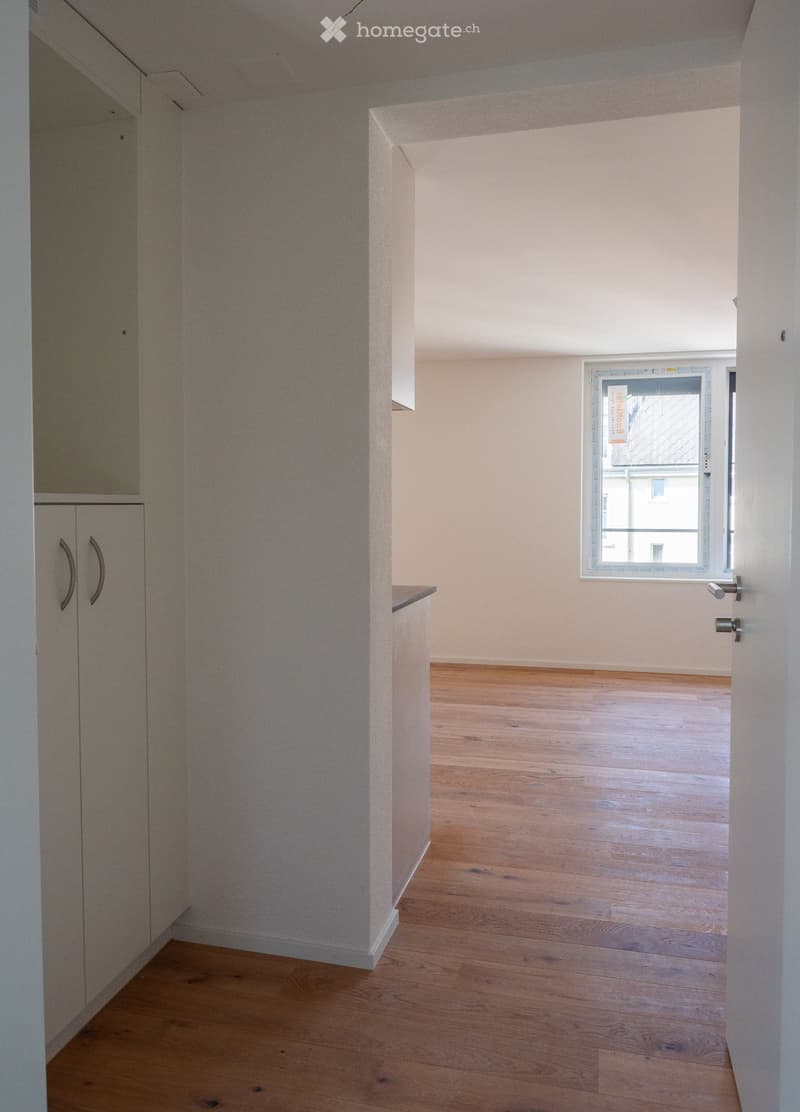 Empty room with wooden flooring, white walls, and a window with a view of a building outside. The room features built-in cabinets with handles on the left side.