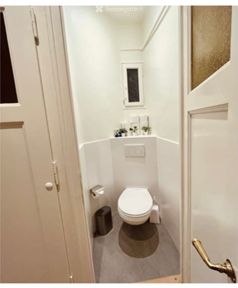 Compact bathroom with white walls, a toilet, a small sink, and a window above the sink. Toilet paper holder and a trash bin are visible.