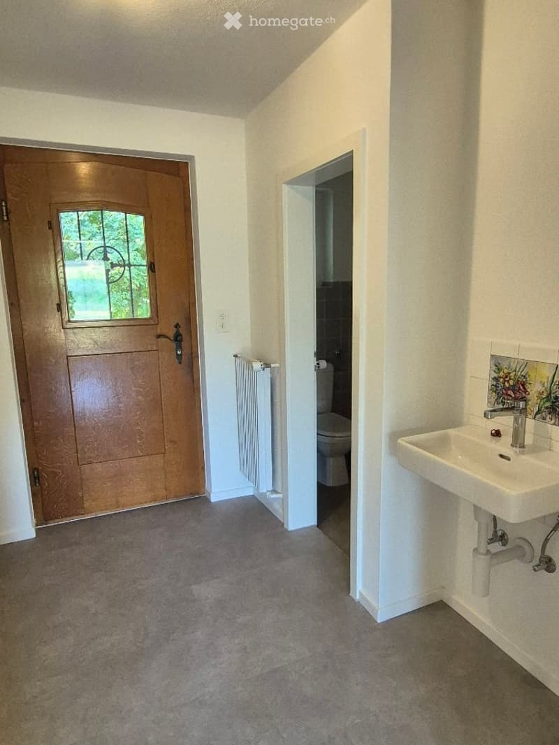 Empty hallway with a wooden front door, a radiator, and an open doorway leading to a bathroom with a sink and toilet.
