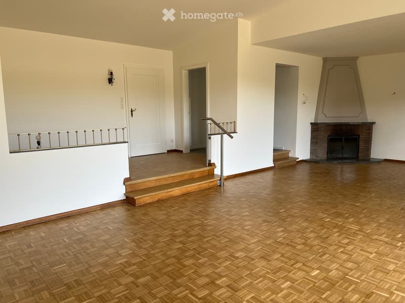 An empty room with wooden flooring, white walls, a staircase leading to another level, and a fireplace with a decorative mantle.