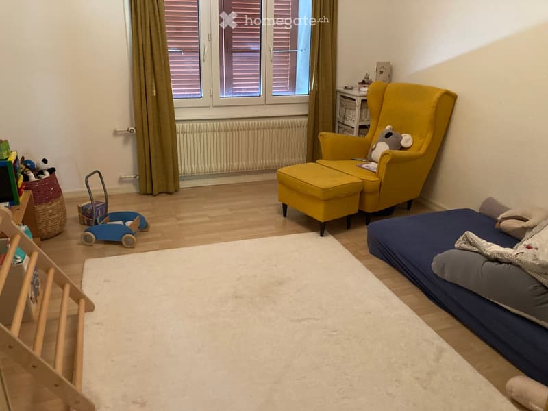 A cozy, well-lit room with a blue bed, yellow armchair, and a small wooden chair. The room features a white rug, a toy car, and a basket with toys.