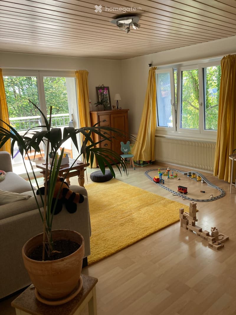 Bright living room with wooden floors, large windows, and a balcony. Features a sofa, coffee table, toy train set, and potted plants.