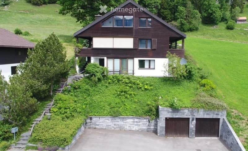 A two-story wooden house with a sloped roof, multiple windows, and a balcony on the upper level. The house is surrounded by greenery and has a stone-walled garage with two doors.