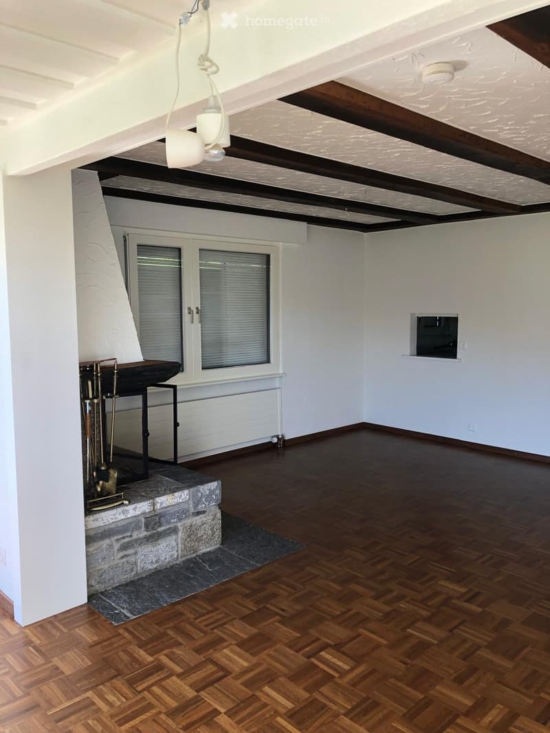 Empty room with wooden flooring, exposed wooden beams on the ceiling, and a stone fireplace with a small table and fireplace tools nearby.