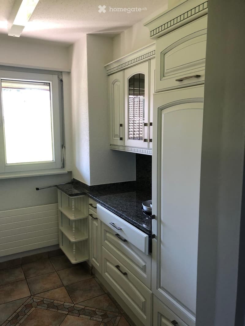 A small kitchen with white cabinets, a dark countertop, and a small bowl on the counter. There is a window with blinds and a small shelf unit next to the window.