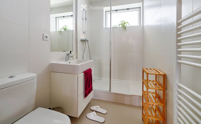 Modern bathroom with white tiles, a sink, mirror, toilet, glass-enclosed shower, a red towel, a wooden shelf, and two white bathroom slippers on the floor.