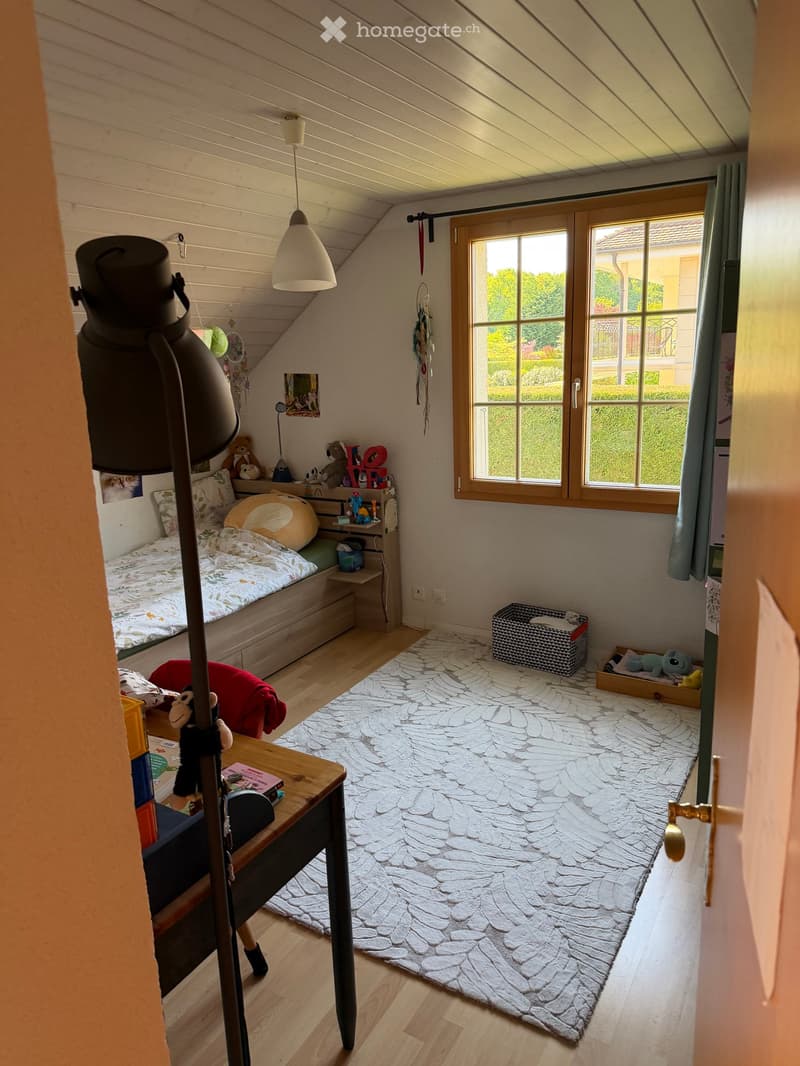 Cozy attic bedroom with slanted wooden ceiling, large window, bed with colorful bedding, small table, and various toys and decorations.