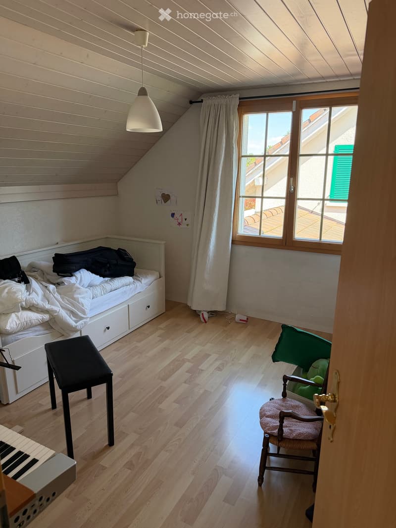 Cozy attic bedroom with wooden floors, slanted ceilings, a built-in bed with drawers, a small black stool, a wooden chair with a cushion, and a hanging lamp.