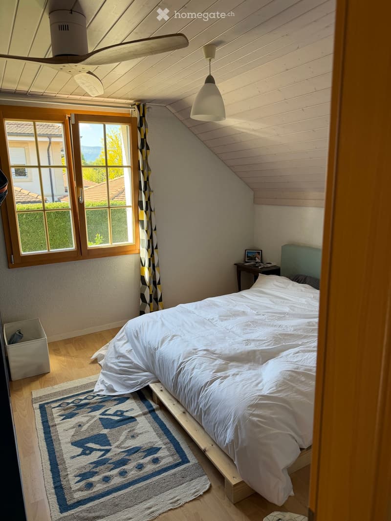 Cozy attic bedroom with wooden slanted ceilings, a bed with white bedding, a nightstand with a lamp, a window with curtains, and a patterned rug.