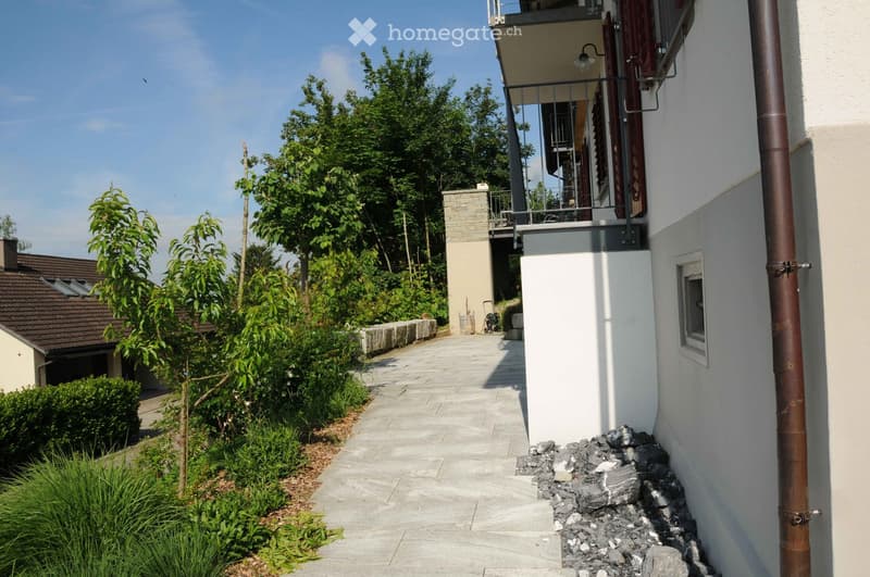 A well-maintained garden area with a paved pathway leading to a modern building. The garden features neatly trimmed bushes, small trees, and a stone wall. A balcony with a metal railing is visible on the building.