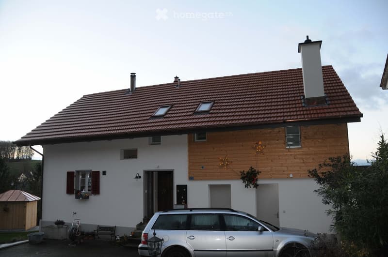 Two-story house with a red-tiled roof, white walls, and wooden siding on the upper level. Features include a chimney, dormer windows, a front entrance, and a parked car in the driveway.