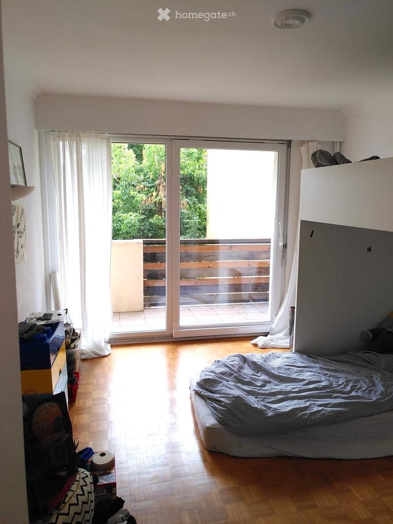 Bright bedroom with wooden flooring, large sliding glass doors leading to a balcony, a bed with striped bedding, a nightstand, and a mirror on the wall.
