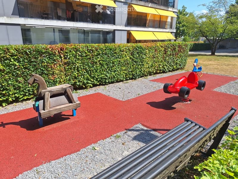 Playground area with a wooden rocking horse, red toy car, and a bench, surrounded by a hedge and a modern building with balconies.