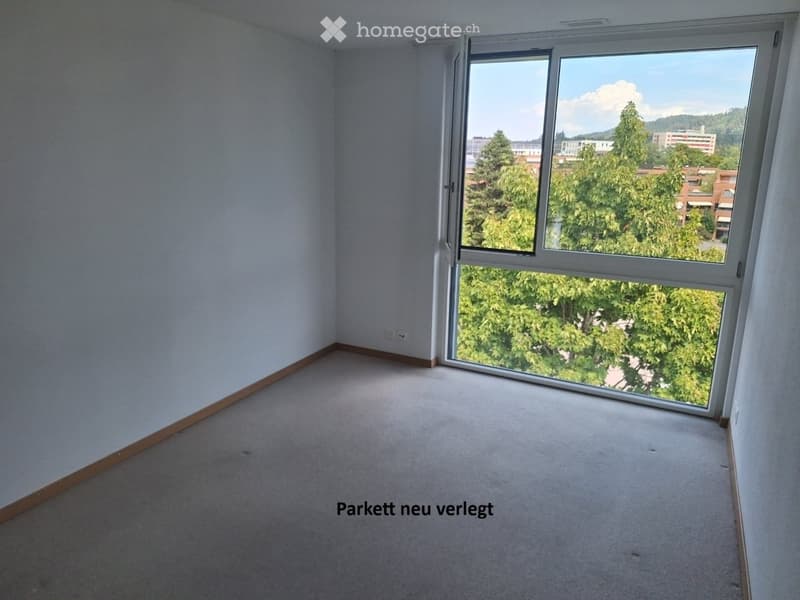 Empty room with light-colored walls, carpeted floor, and large window offering a view of trees and buildings.