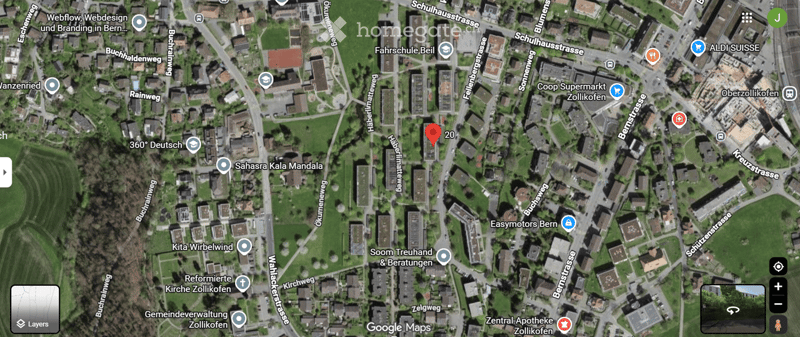 Aerial view of a residential area with multiple buildings, streets, and green spaces. Marked location at the center indicates a specific point of interest.