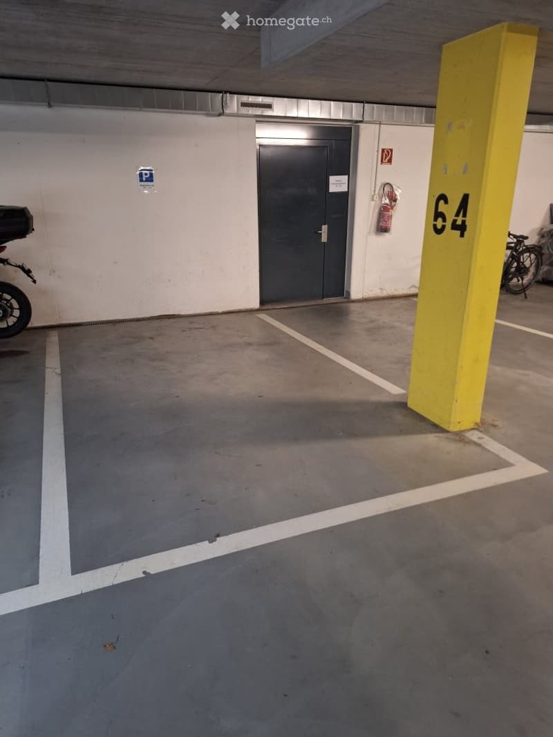 Underground parking area with marked parking spaces, a yellow pillar marked '64', a closed metal door, and a fire extinguisher mounted on the wall.