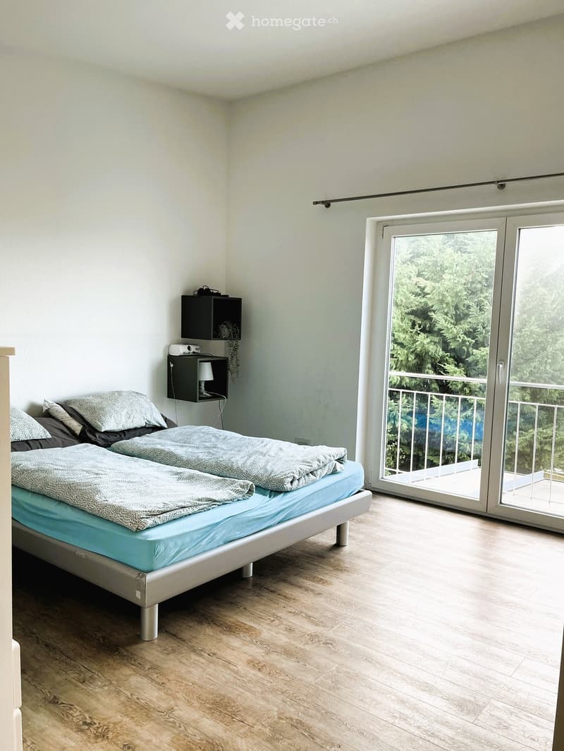 Modern bedroom with a double bed, nightstand, lamp, and a sliding glass door leading to a balcony with a view of greenery.