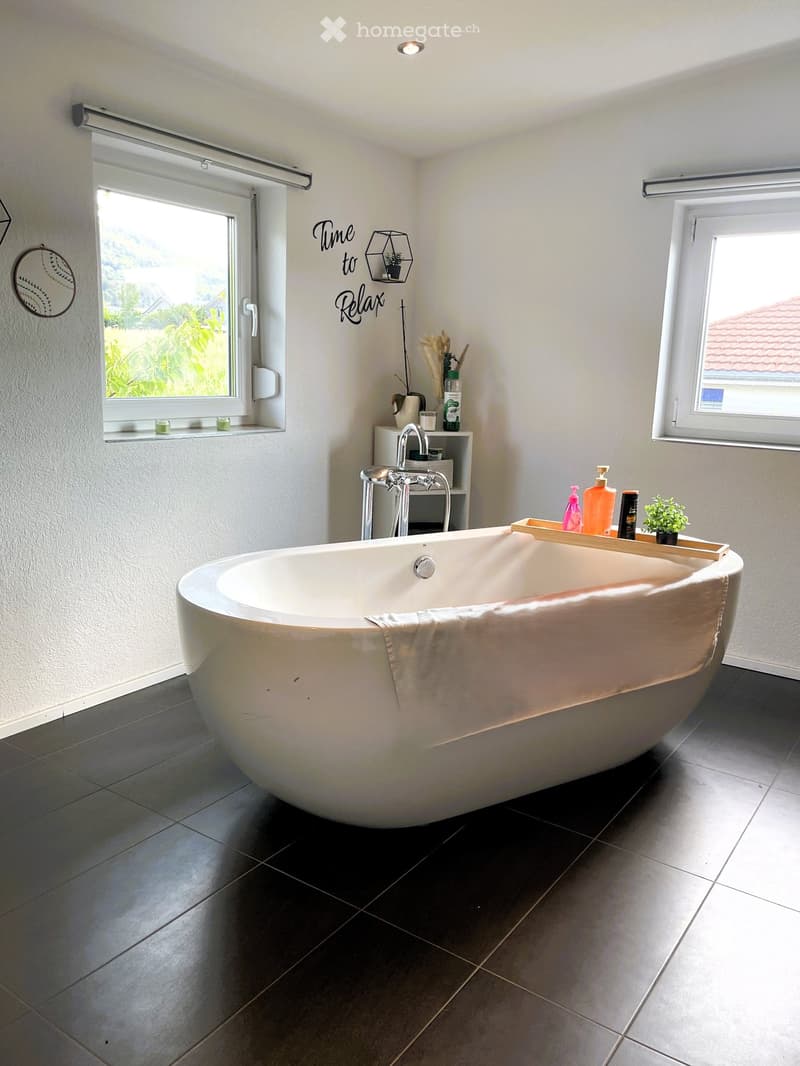 Modern bathroom with a large, freestanding bathtub, a towel draped over the edge, a small shelf with toiletries, a decorative mirror, and candles on the windowsill.