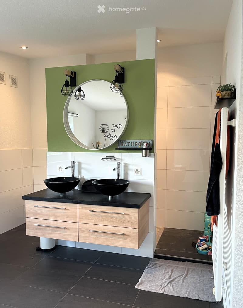 Modern bathroom with a double sink vanity, black countertop, wooden cabinet, large round mirror, wall-mounted lights, and a towel on the floor.