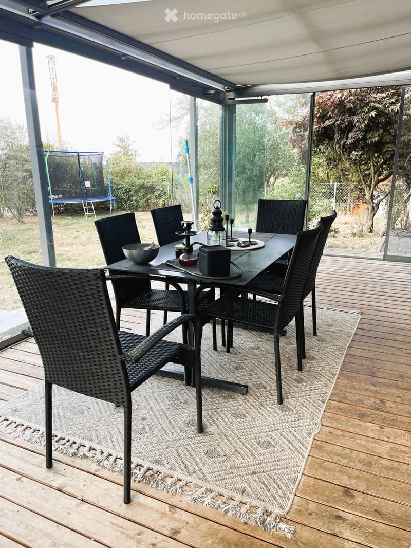Wooden deck with a dining set including a black table and six black wicker chairs, placed on a light-colored rug with fringe edges. The area is enclosed by large glass panels, with a trampoline visible in the background.