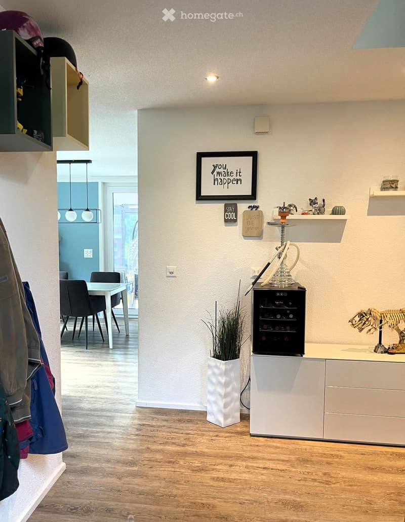 A modern, well-lit room with wooden flooring, a white table and chairs, a wine cooler, shelves with decorative items, and a potted plant. The room features a coat rack with jackets and a framed motivational quote on the wall.