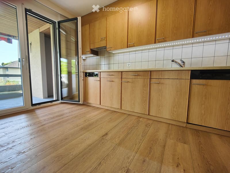 Modern kitchen with light wooden cabinets, a sleek countertop, and a built-in dishwasher. The space features a polished wooden floor and a large sliding glass door leading to an outdoor area.