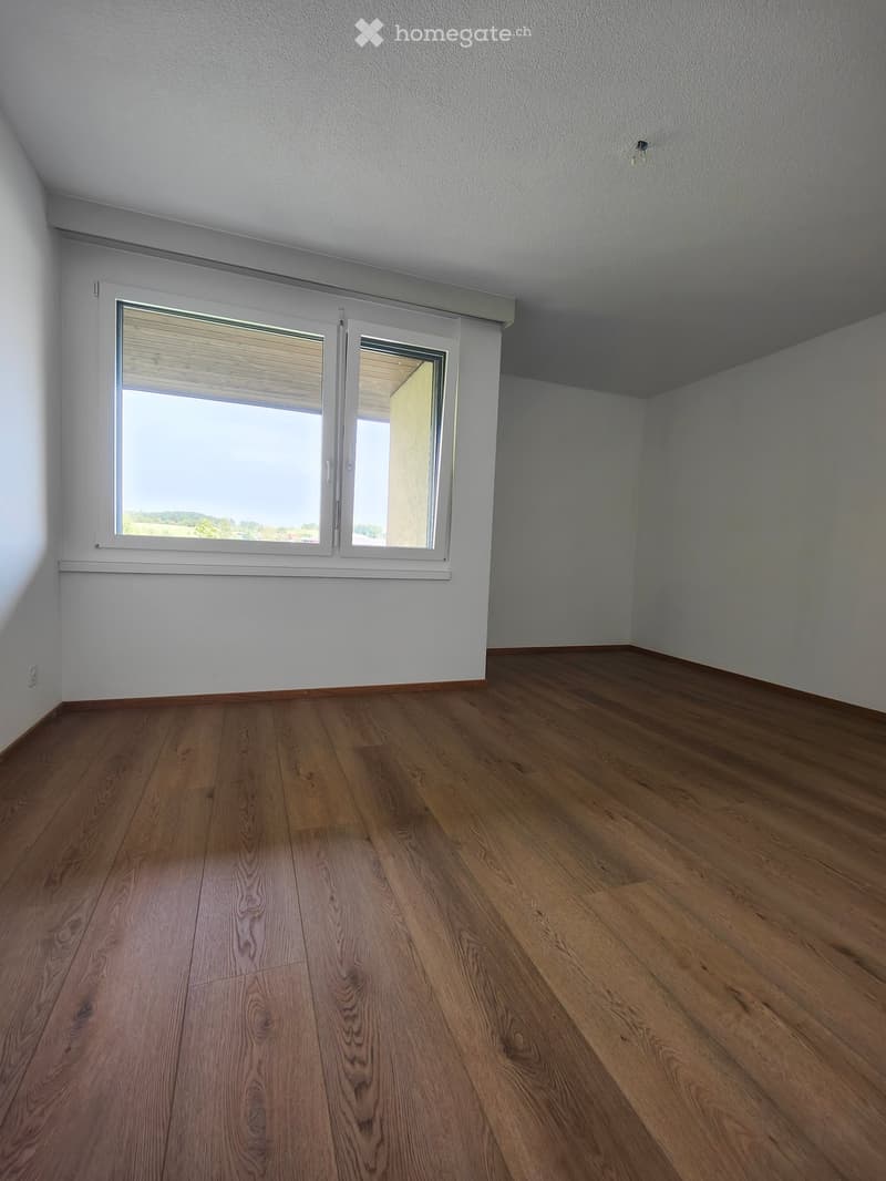 Empty room with wooden flooring and white walls, featuring a large window with blinds and a view of the outdoors.