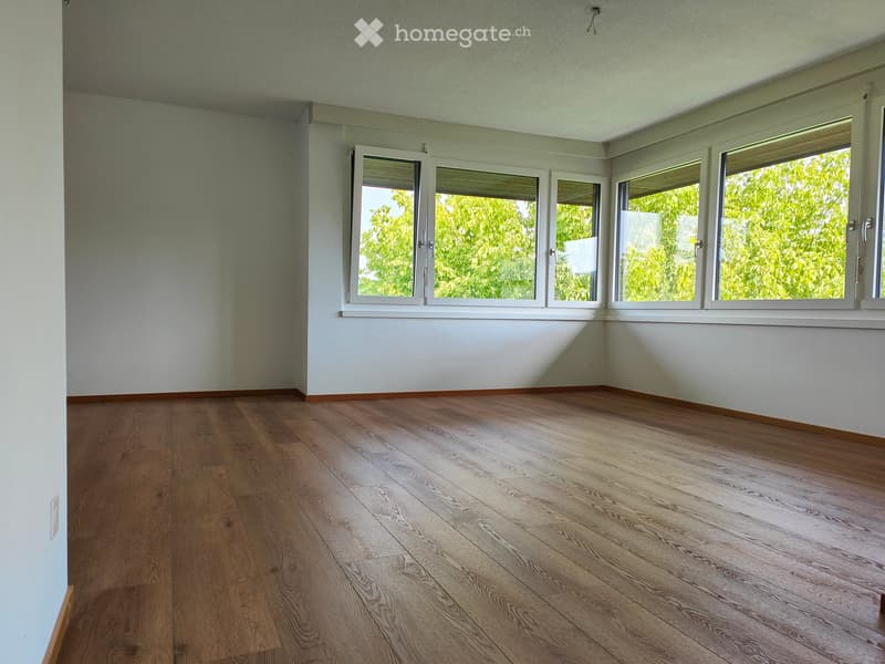 Empty room with wooden flooring and large windows, featuring light-colored walls and a clean, minimalist design.